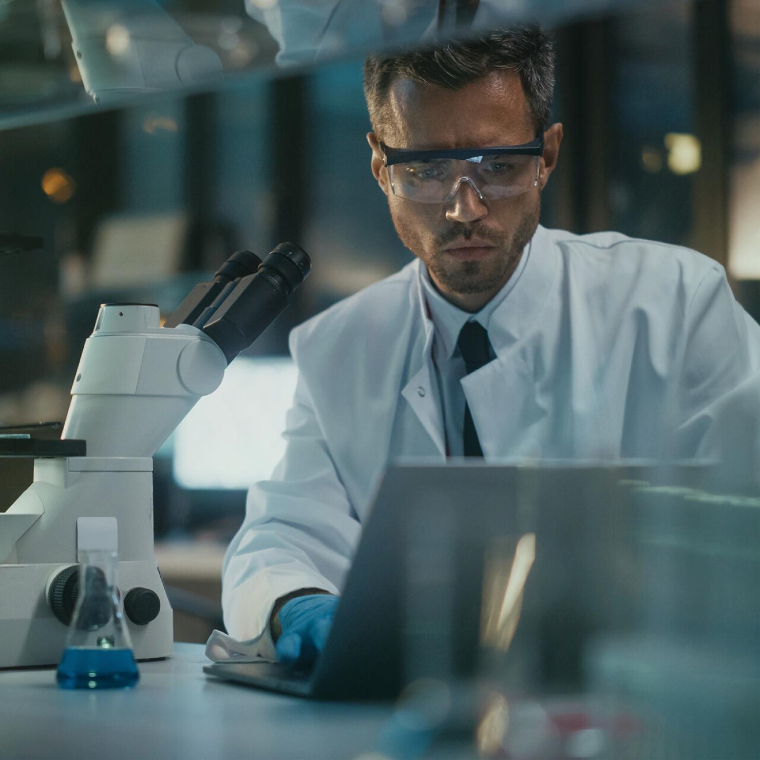 Medical technician in lab coat working on laptop and microscope.